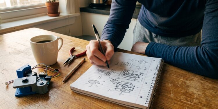 Inventor sketching exploded mechanical diagrams in a notebook at a kitchen table, prototype with wires beside coffee mug, natural light
