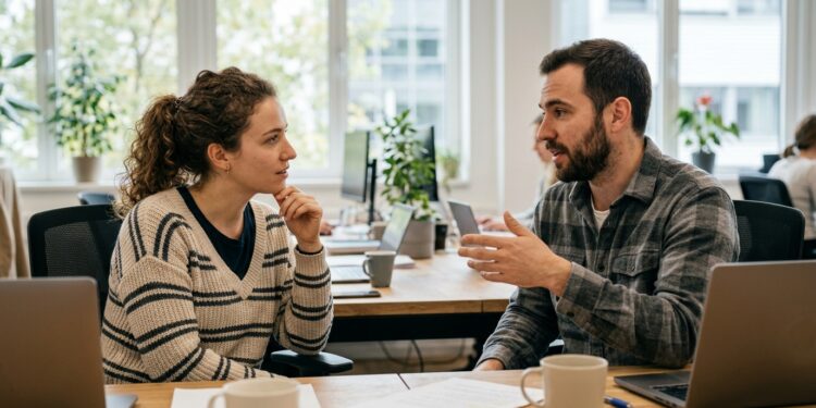Two colleagues in an office, one leaning in and listening attentively while the other speaks